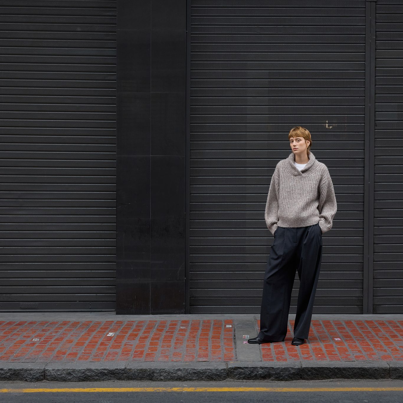 A person stands on a red brick sidewalk in front of dark gray metal storefront shutters, wearing a light beige/gray knit cowl-neck sweater and dark wide-leg pants.