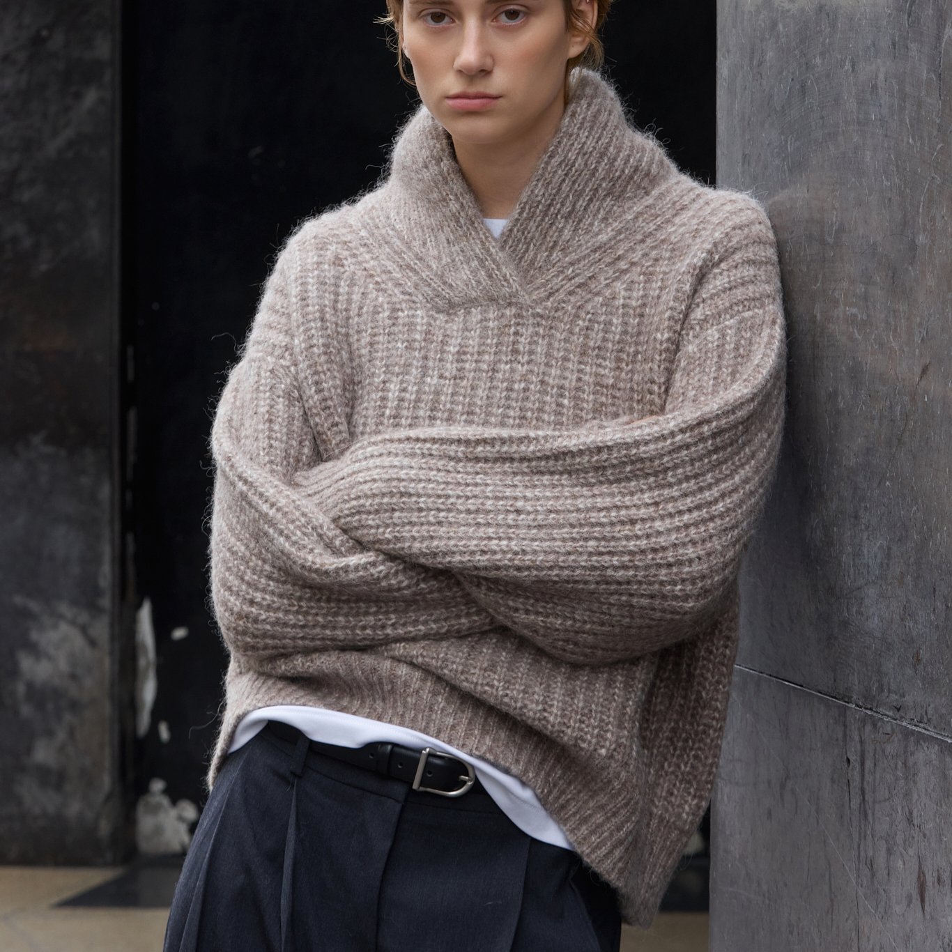 A model wearing a chunky, oversized ribbed knit cowl-neck sweater in a taupe-beige color, styled with dark trousers and a white undershirt, leaning against a dark textured wall.