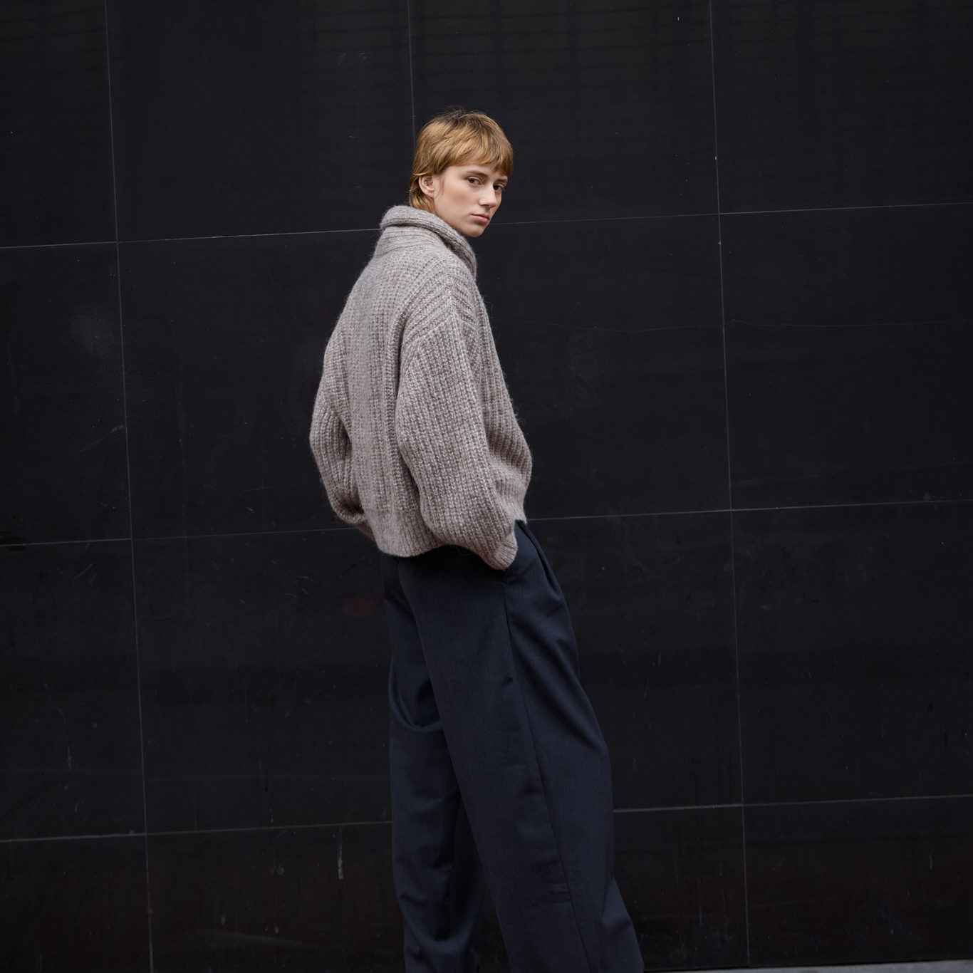 A model wearing a chunky knit beige sweater and dark navy wide-leg trousers, standing against a dark tiled wall.
