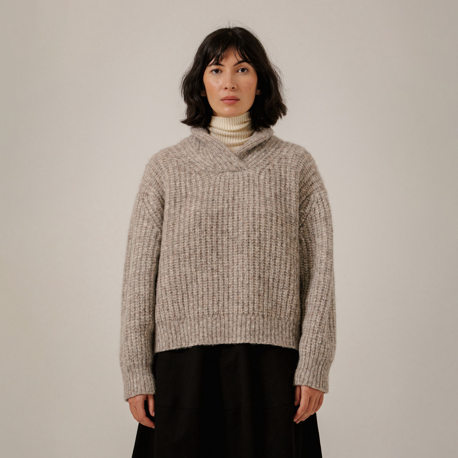 A woman wearing a chunky knit wrap-neck sweater in a heathered taupe-beige color, layered over a cream turtleneck and a black skirt, standing against a plain light gray studio background.