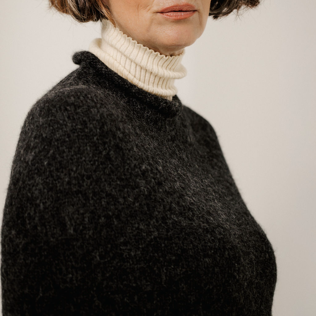 Close-up of a person wearing a dark charcoal fuzzy poncho sweater over a cream ribbed turtleneck, against a plain light background.