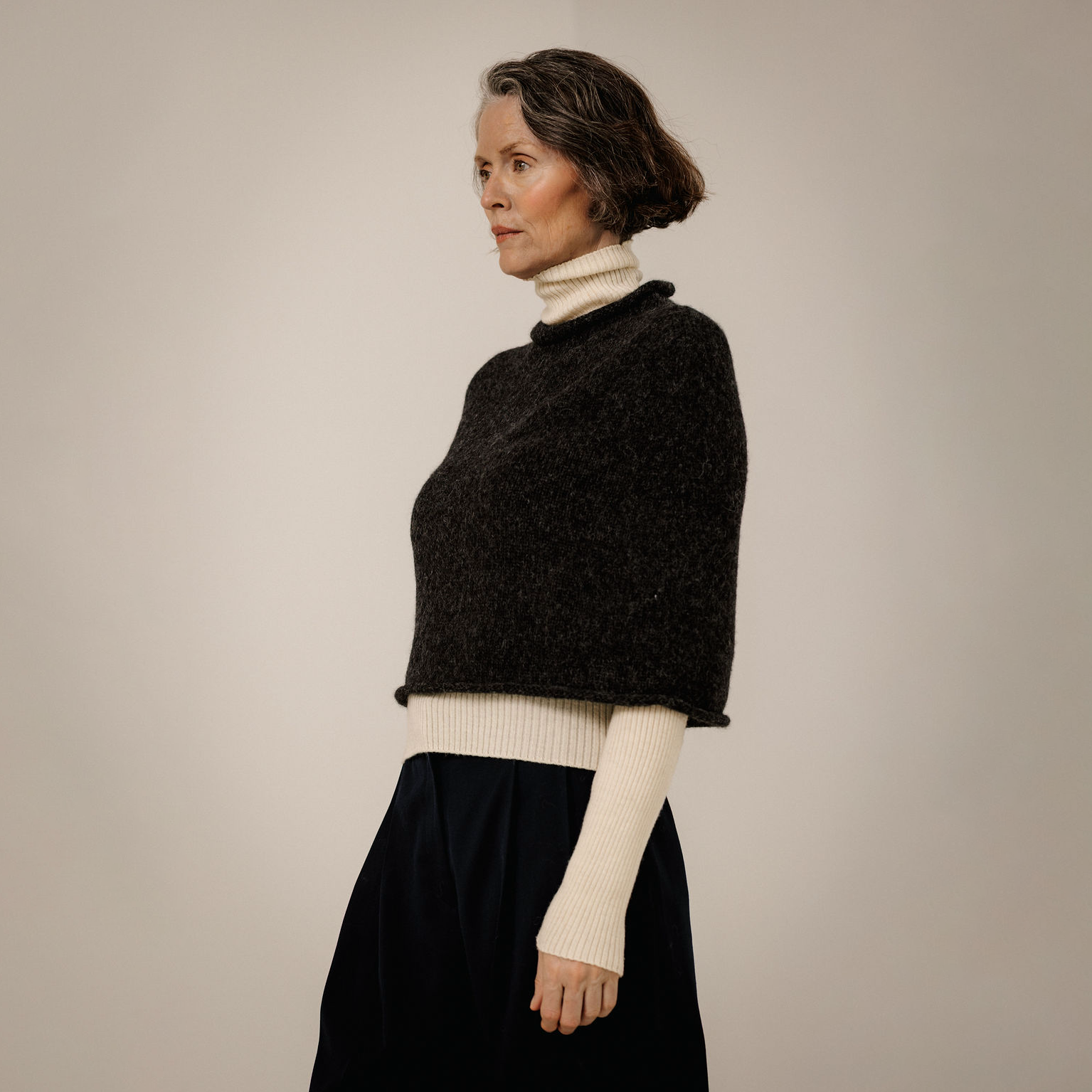A woman in profile wearing a dark charcoal knit cropped poncho over a cream ribbed turtleneck, paired with a dark skirt, against a plain light beige studio background.