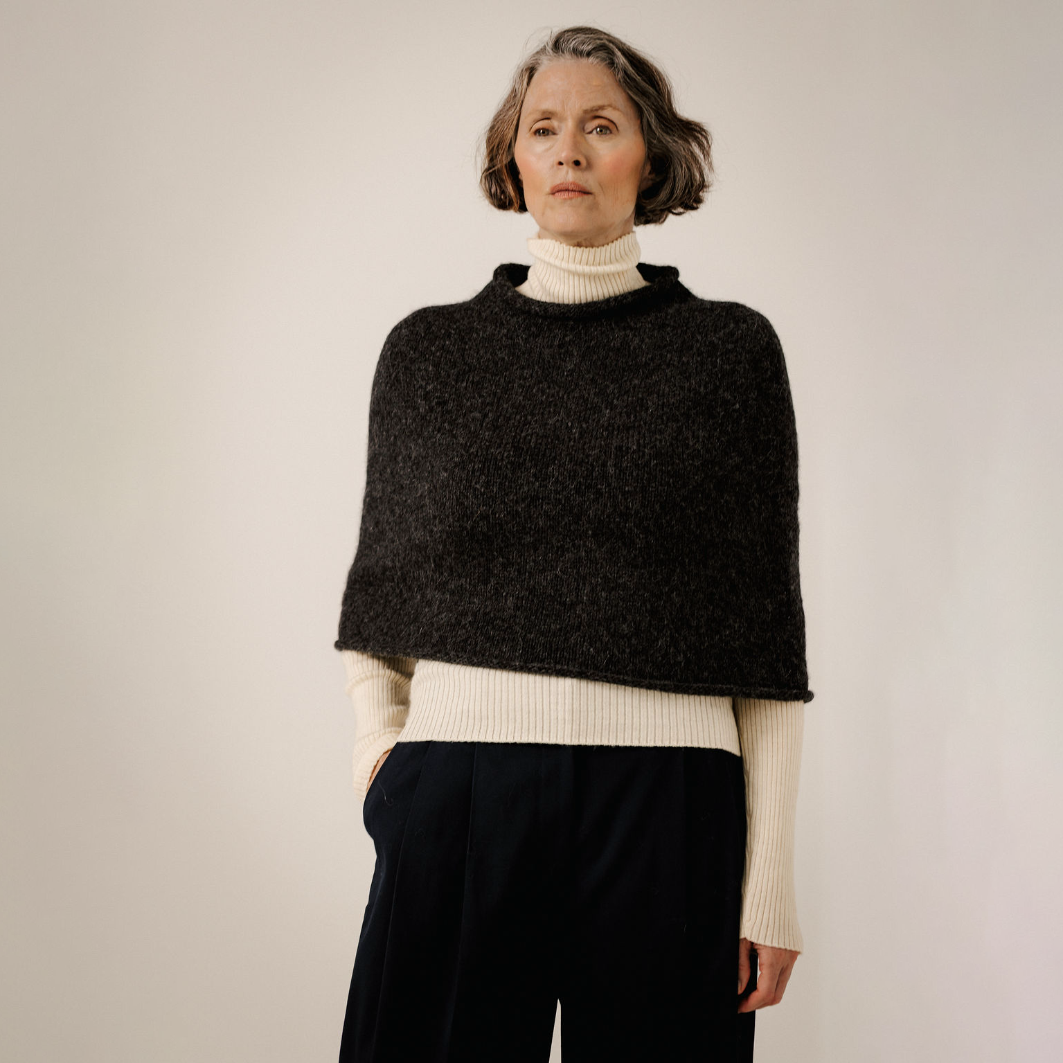 A woman modeling a dark charcoal knit cape sweater over a cream ribbed turtleneck and dark trousers, photographed against a plain light beige studio background.