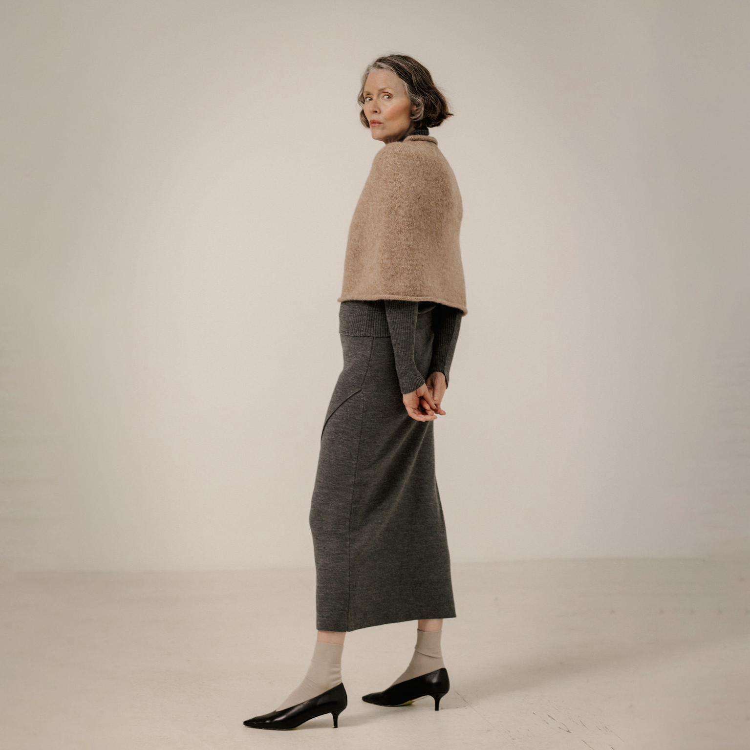 A woman stands in profile wearing a beige cropped knit top, a dark gray midi skirt, light socks, and black pointed-toe heels against a plain light studio background.