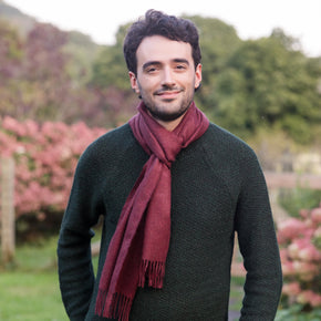 A person wearing a red solid color baby alpaca scarf with tassels, standing outdoors with greenery and pink flowers in the background.