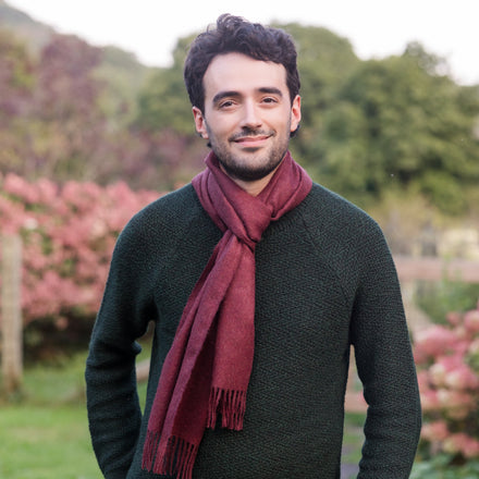 A person wearing a red solid color baby alpaca scarf with tassels, standing outdoors with greenery and pink flowers in the background.