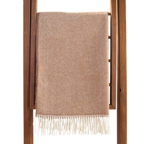 Fawn woven blanket with white fringe draped over a wooden ladder on a white background