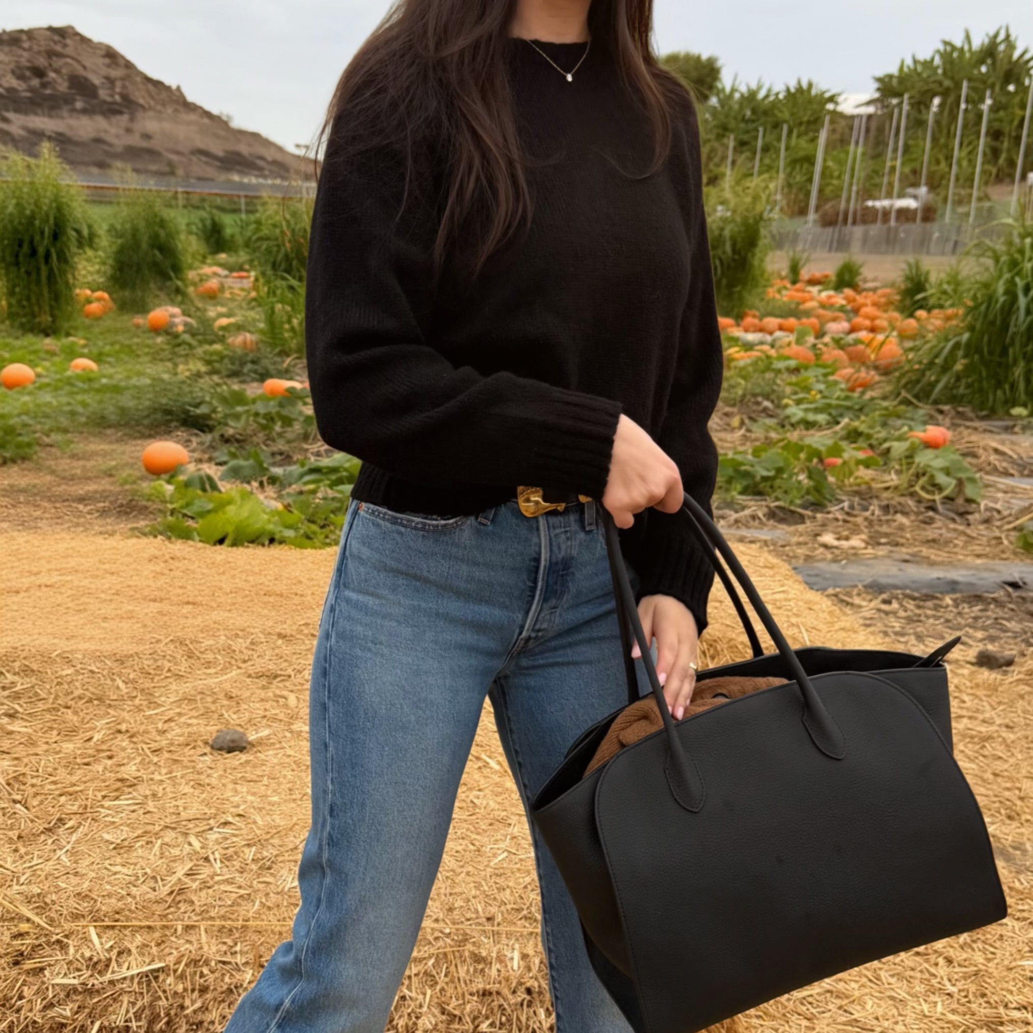 A woman standing outdoors in a pumpkin patch wearing a black sweater, blue jeans, and black boots, while holding a large black tote bag.