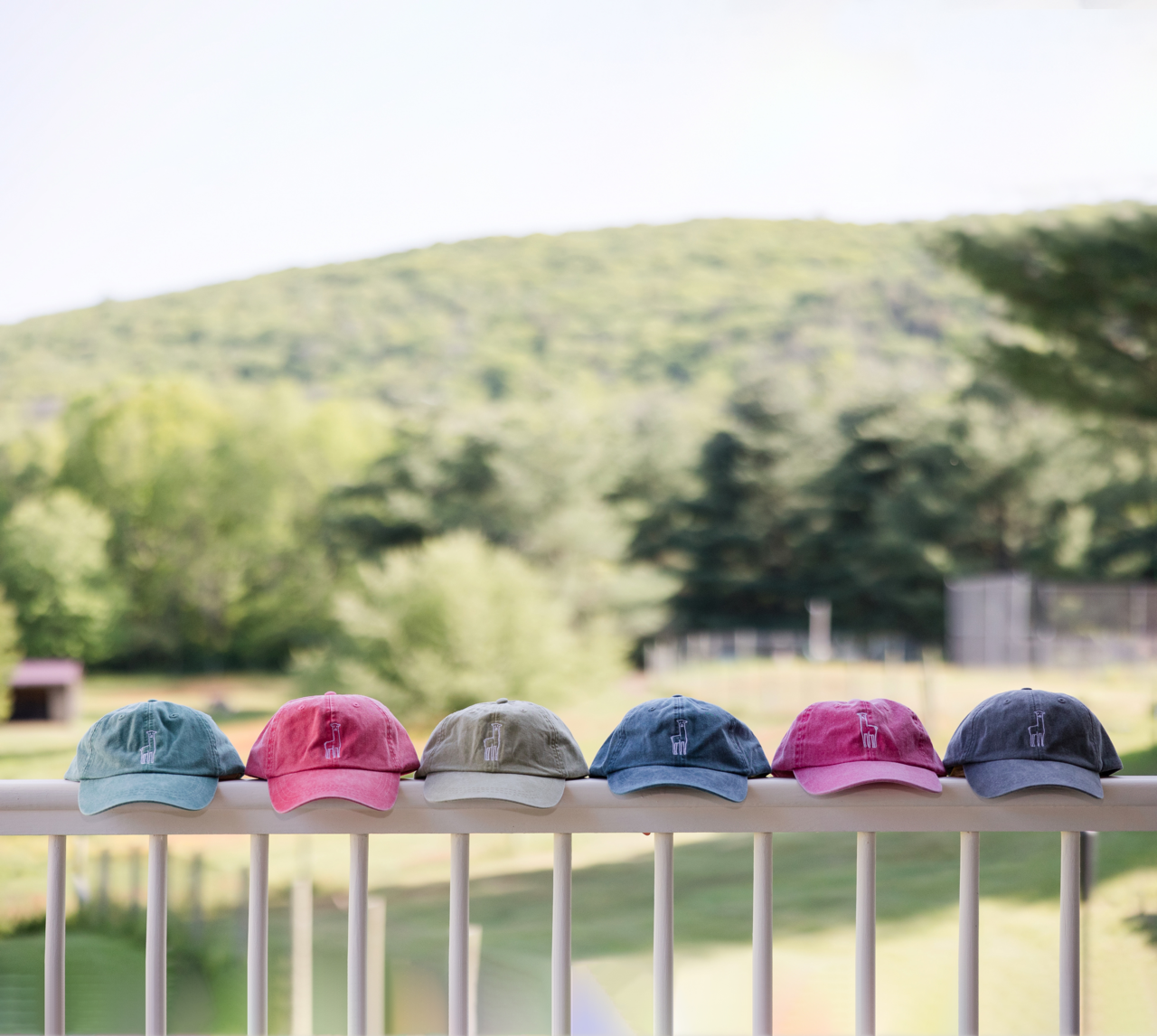 6 Fluff Alpaca Embroidered Baseball Hats sitting in a row on a white railing against a backround of farm fields and rolling green hills.