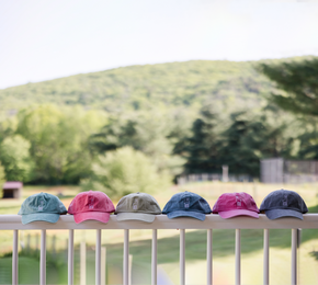 6 Fluff Alpaca Embroidered Baseball Hats sitting in a row on a white railing against a backround of farm fields and rolling green hills.