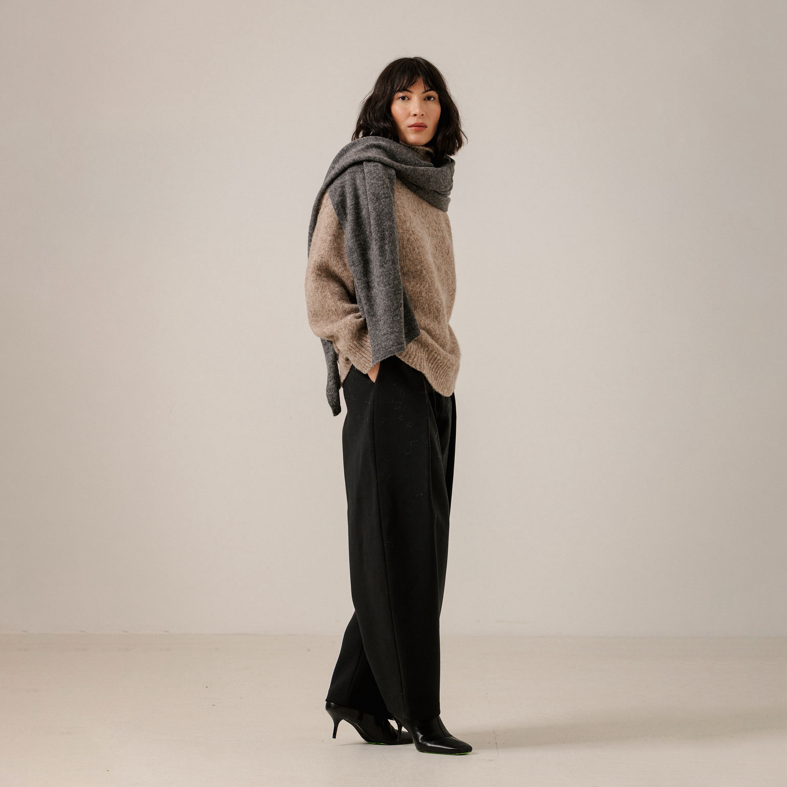 A woman stands in profile wearing a chunky knit beige sweater, a thick gray scarf, wide-leg black pants, and black pointed heels against a plain light gray studio background.
