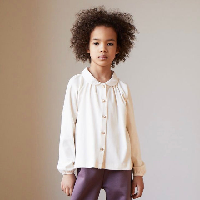 A girl wears a white Oeuf Kid's Long Sleeve Blouse and purple pants in front of a neutral background.