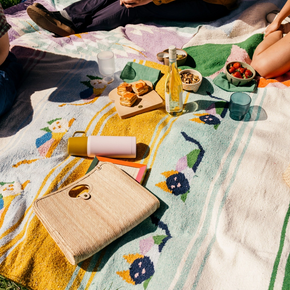 People sitting on a colorful blanket with food and drinks in a natural setting