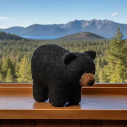 Black bear felted toy on a windowsill with a scenic view of mountains and trees outside.