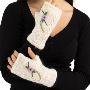 White knitted wrist warmers with floral embroidery held by a person wearing a black shirt.