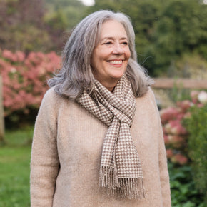 An older woman with grey hair is smiling, wearing a beige knit sweater and a beige and white houndstooth patterned scarf with tassels.