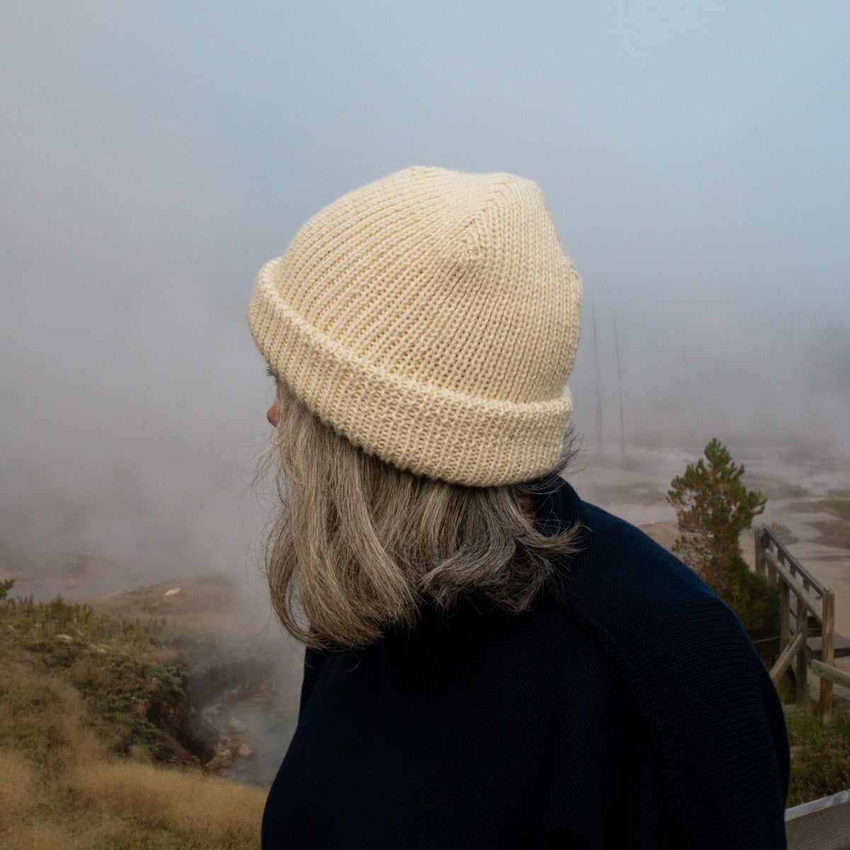A person seen from the side wearing a cream-colored knit beanie hat, standing outdoors in a misty, foggy landscape.