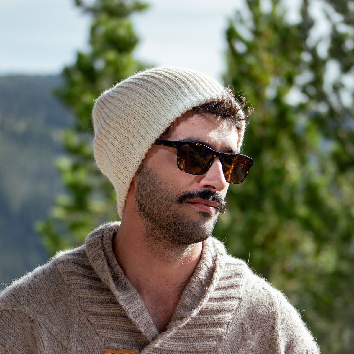 A man wearing a cream ribbed knit beanie, tortoiseshell sunglasses, and a beige knit sweater, photographed outdoors with blurred green trees in the background.