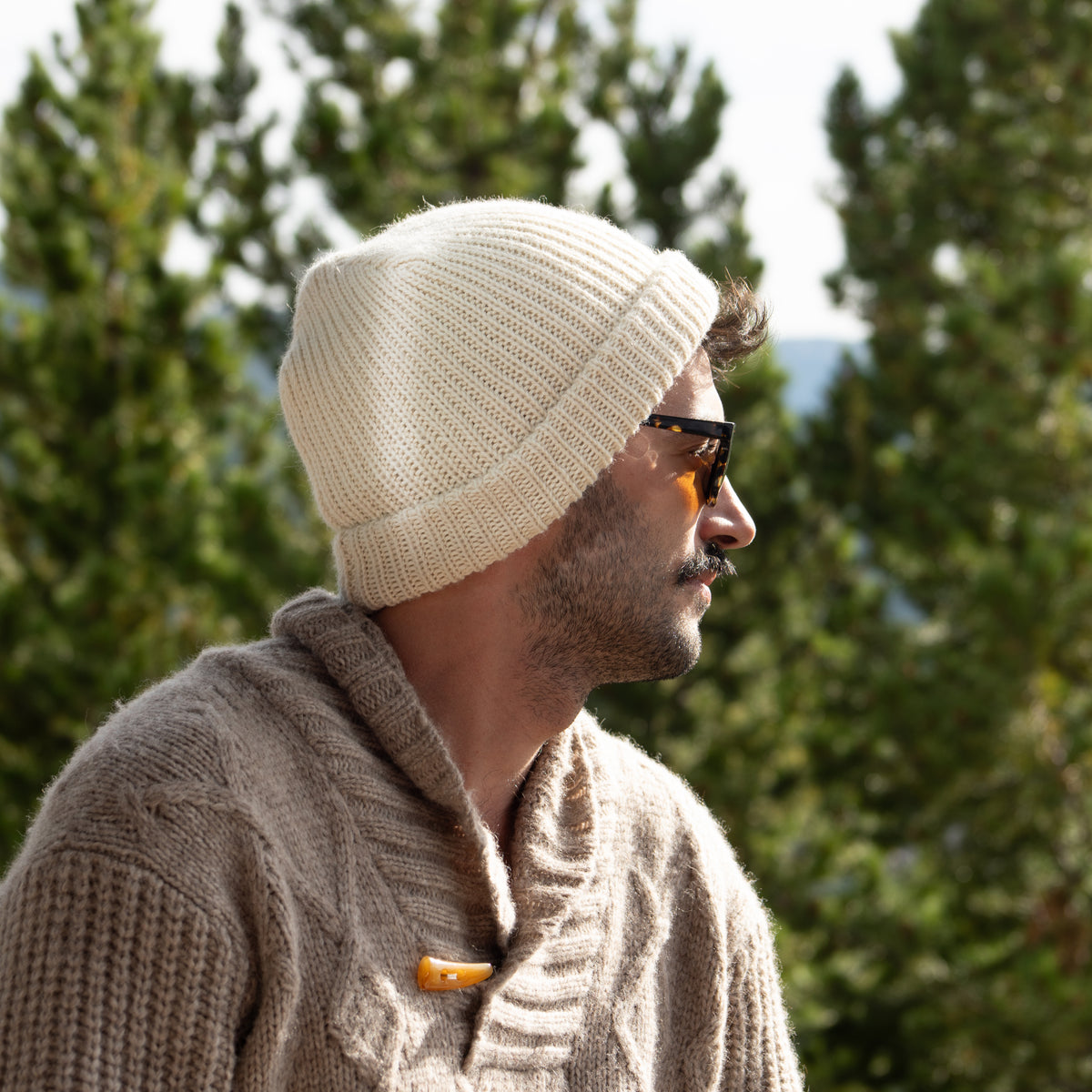 A man in profile wearing a cream ribbed knit beanie and a taupe cable-knit sweater, with green trees blurred in the background.