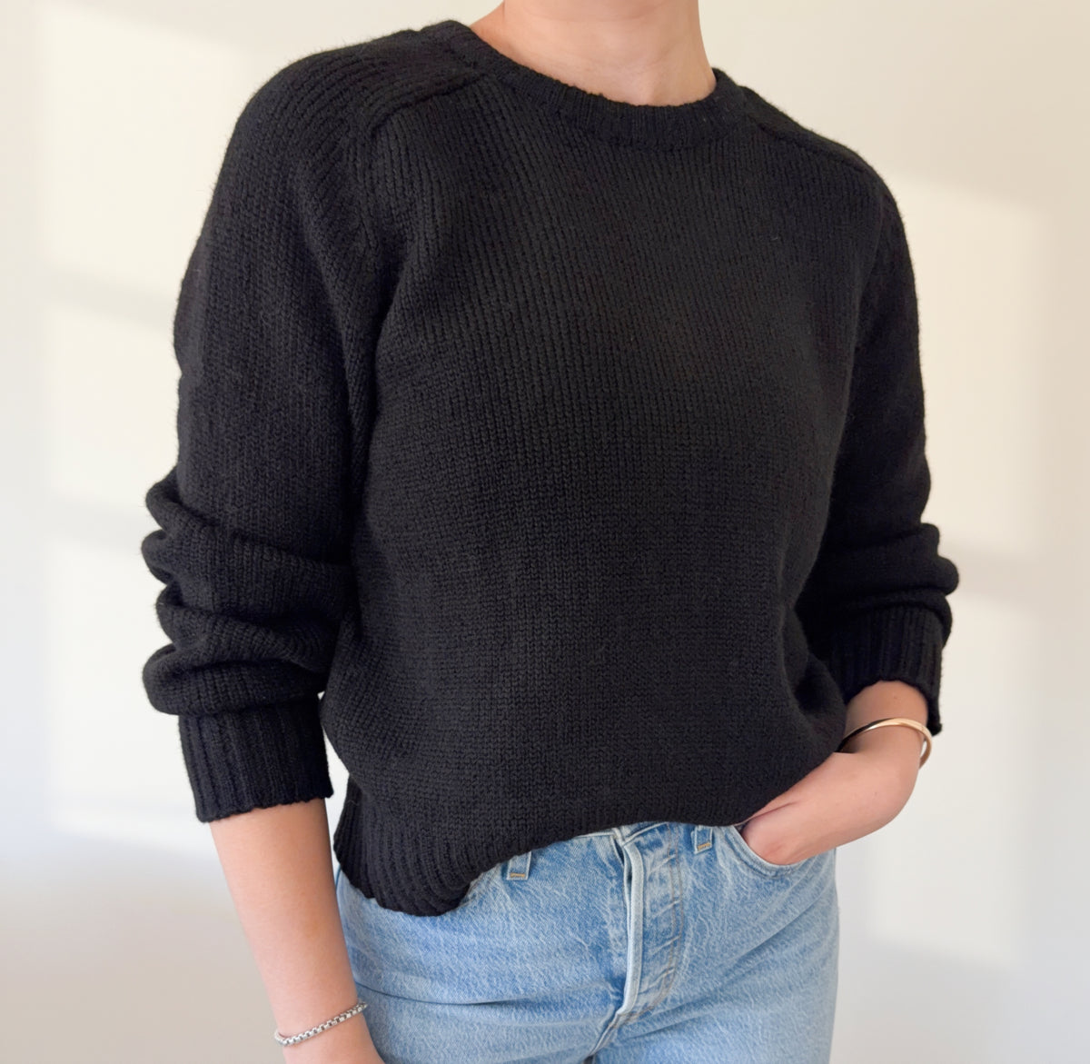A person wearing a black ribbed knit crewneck sweater with cuffed sleeves, paired with light blue jeans, standing against a plain off-white wall.