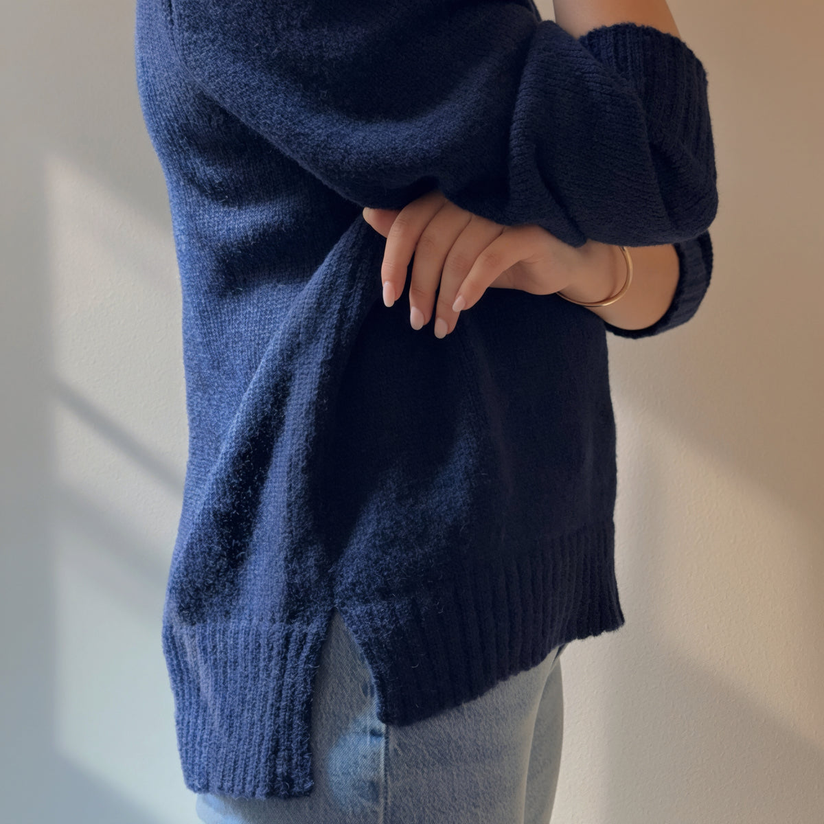A person wearing a dark navy blue fuzzy knit sweater with ribbed cuffs and hem, paired with light blue jeans, shown from the side against a neutral beige wall.