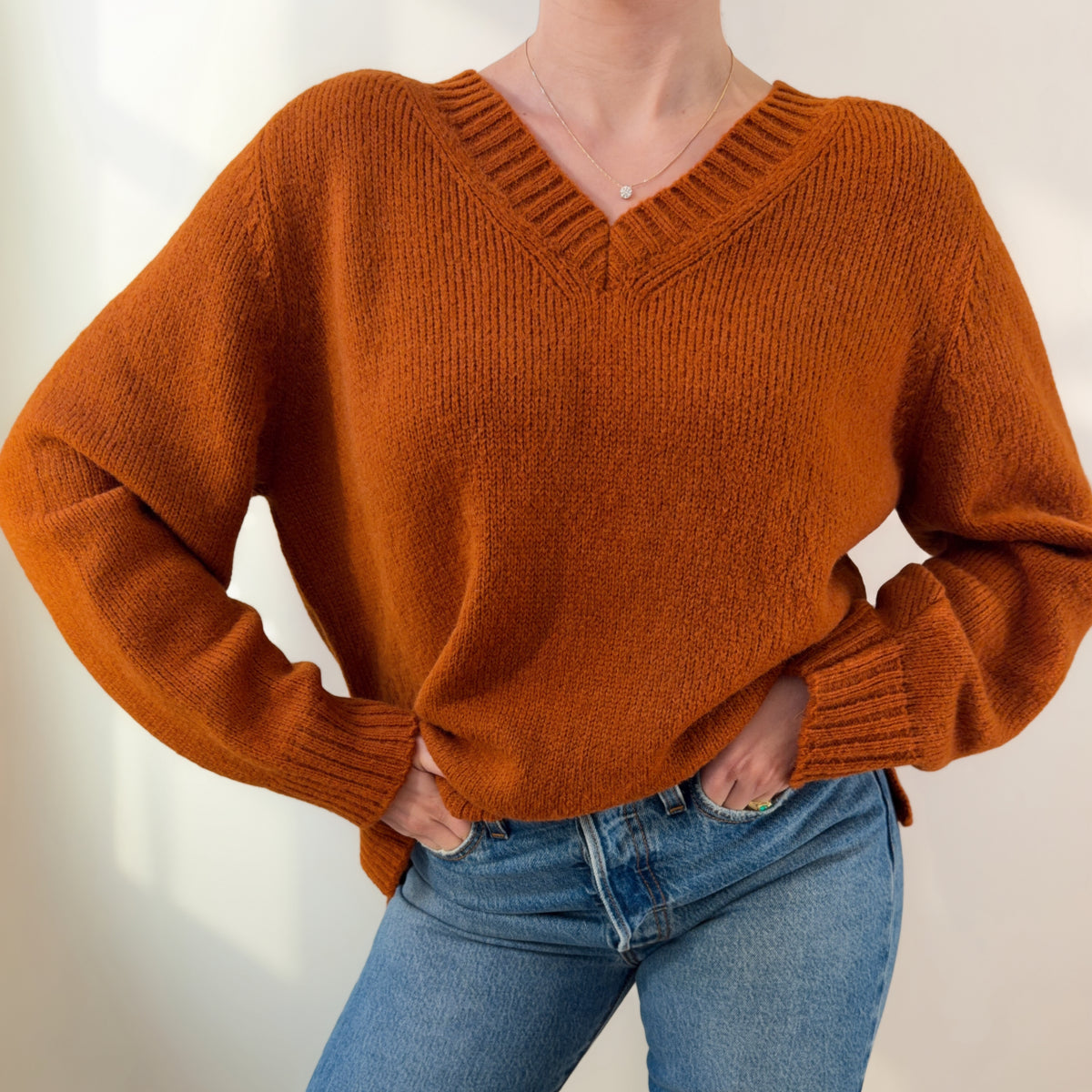A person wearing a chunky knit V-neck sweater in a warm burnt orange color, paired with blue jeans, shown from the neck down against a plain light background.
