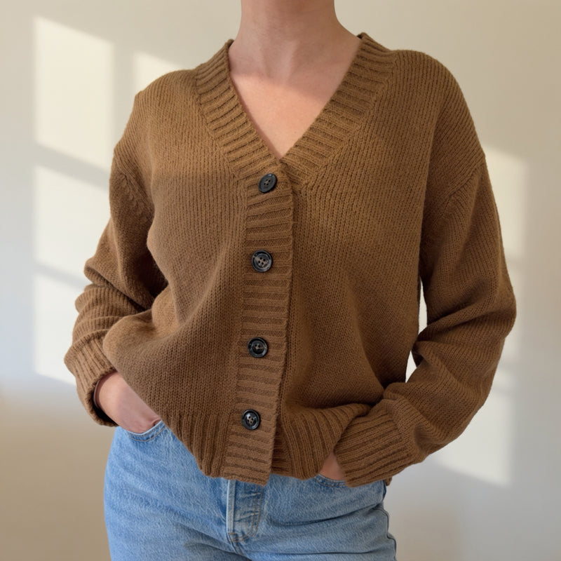 A person wearing a brown ribbed knit cardigan with dark buttons and light blue jeans, standing against a softly lit off-white wall.