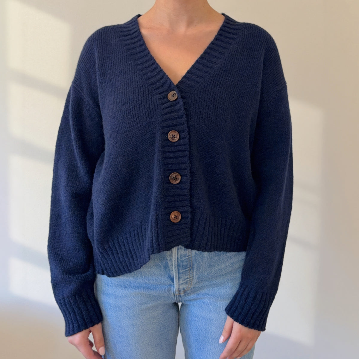 A person wearing a navy blue knit cardigan sweater with brown buttons, paired with light blue jeans, standing against a plain light wall.