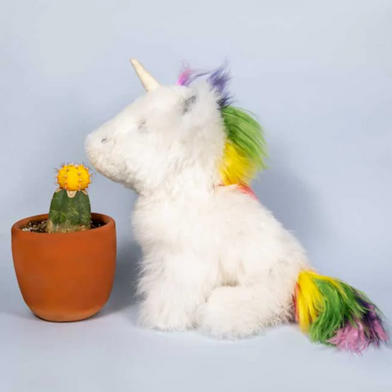 Stuffed unicorn with a rainbow mane and tail next to a potted cactus on a light gray background