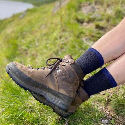 Brown hiking boot and blue socks on a person sitting on a grassy hill with a scenic background.
