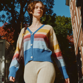 Woman wearing a colorful striped sweater standing outdoors with trees and a building in the background.