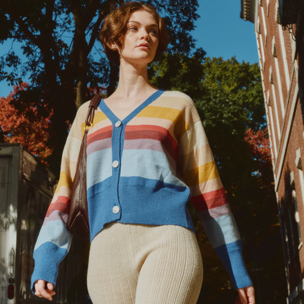 Woman wearing a colorful striped sweater standing outdoors with trees and a building in the background.