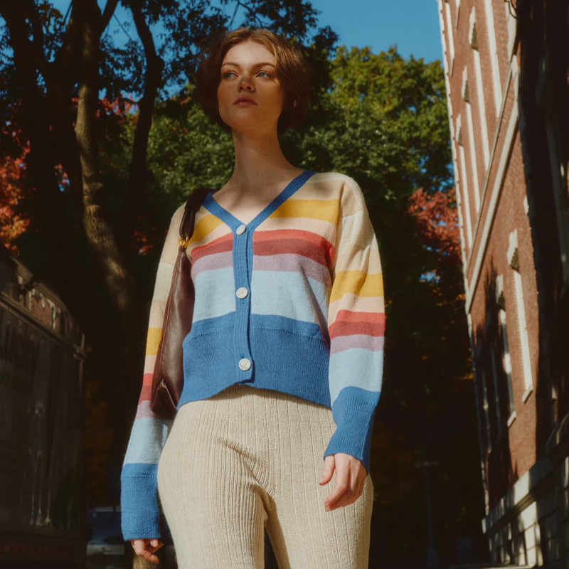 Woman wearing a colorful striped sweater and beige pants standing on a street with trees and buildings in the background.