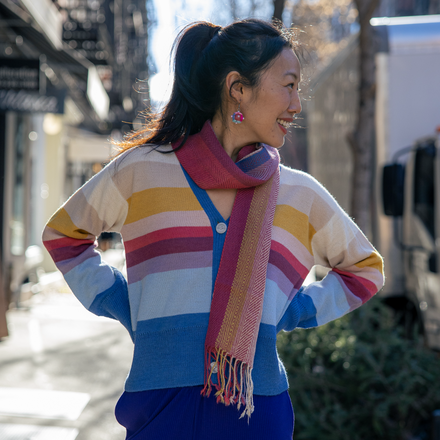 Woman wearing a colorful striped sweater and blue pants with a pink scarf, standing outdoors.