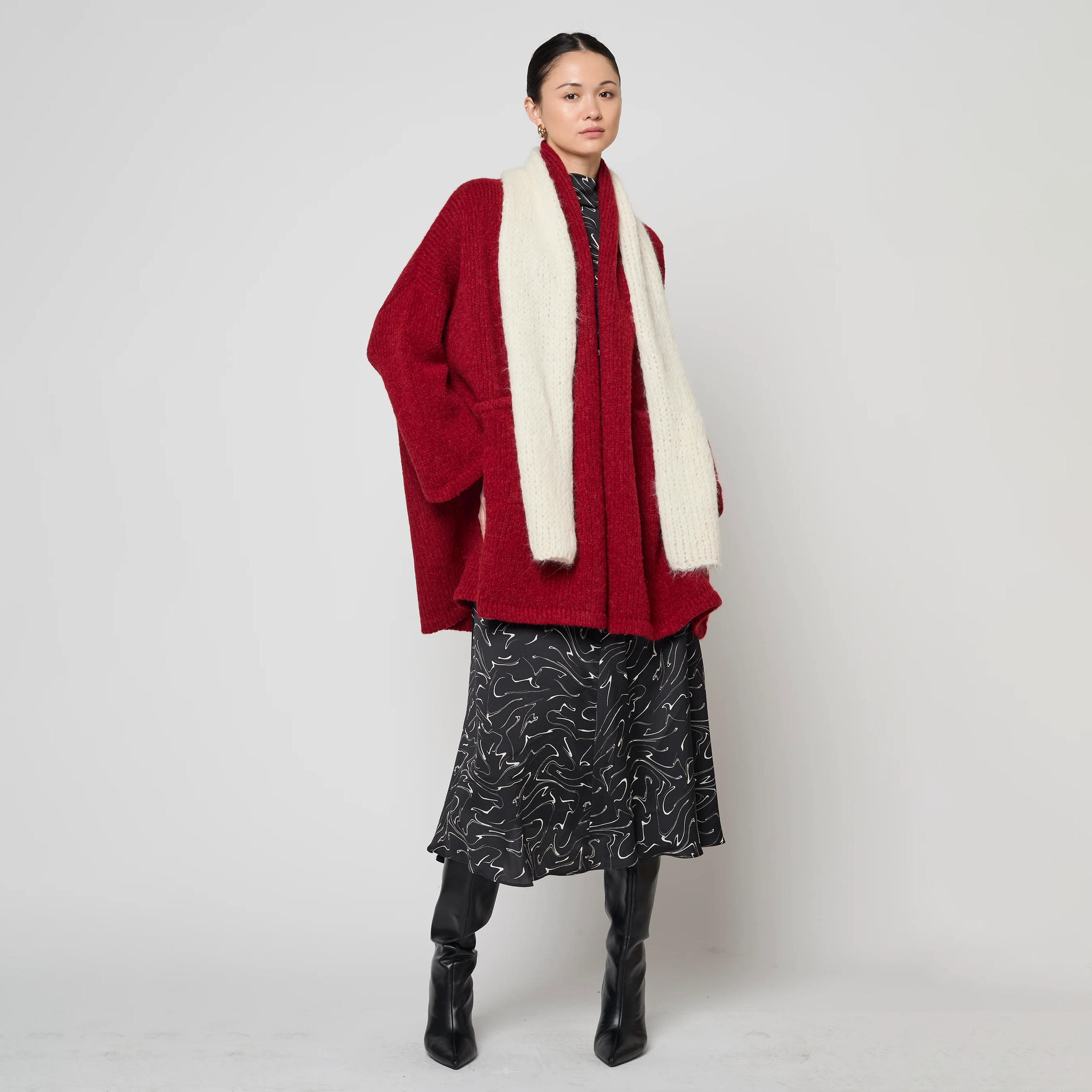Person wearing a red cardigan, white scarf, black patterned skirt, and black boots on a white background
