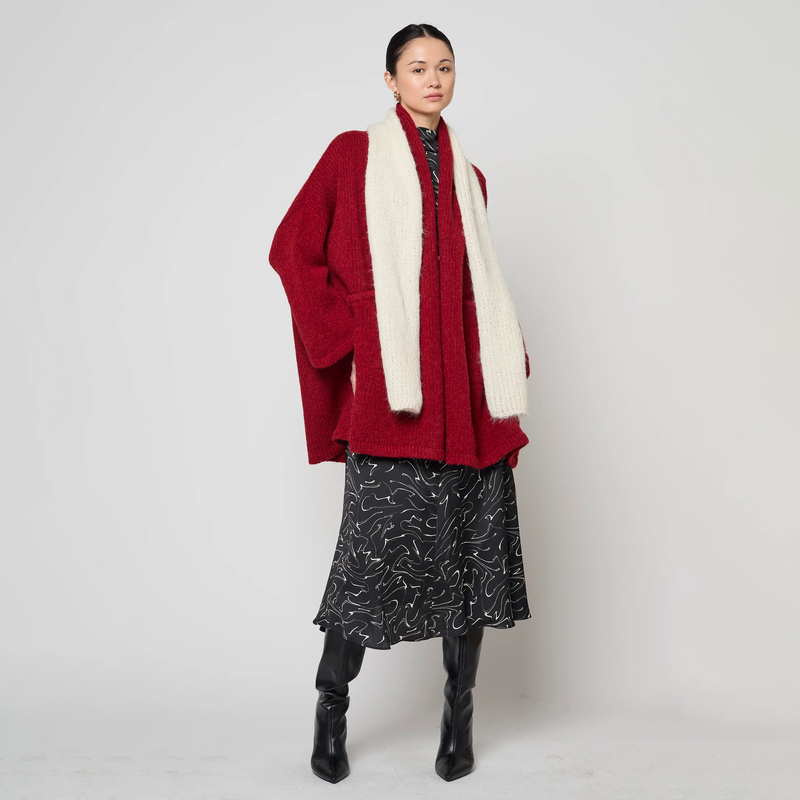 Person wearing a red cardigan, white scarf, black patterned skirt, and black boots on a white background