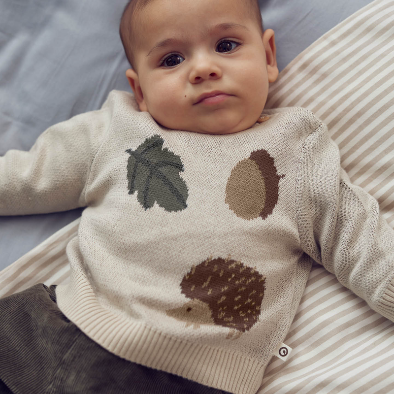 Baby wearing a beige sweater with nature-themed designs on a striped surface