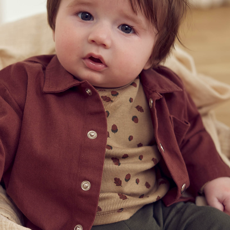 Baby wearing a brown jacket over a beige shirt with acorn pattern, sitting on a beige surface.