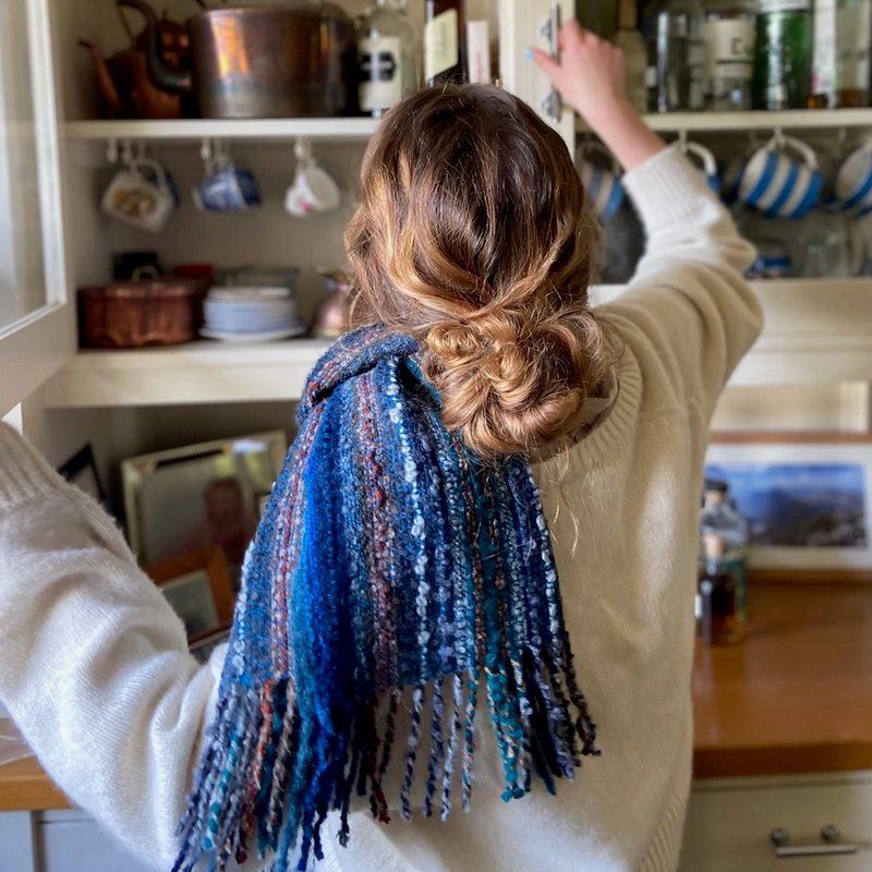 Person with a blue scarf reaching into a kitchen cabinet filled with bottles and cookware.