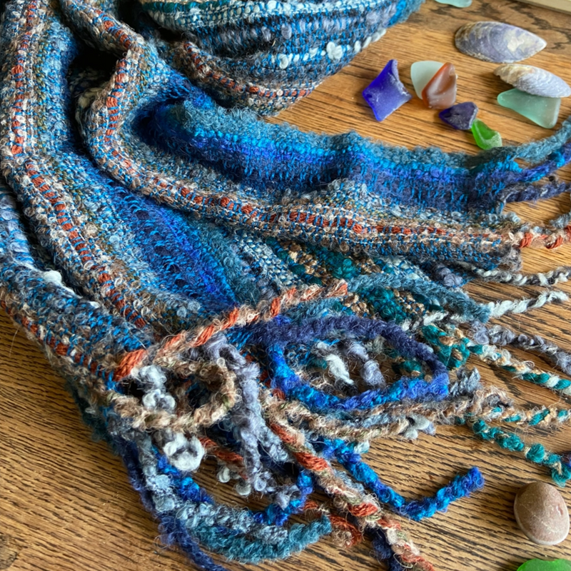 Textured blue scarf on a wooden surface with seashells and sea glass.