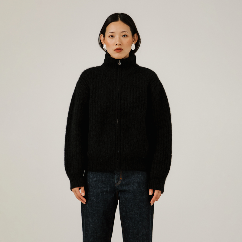Person wearing a black turtleneck zip up rib knit sweater and dark jeans against a plain background