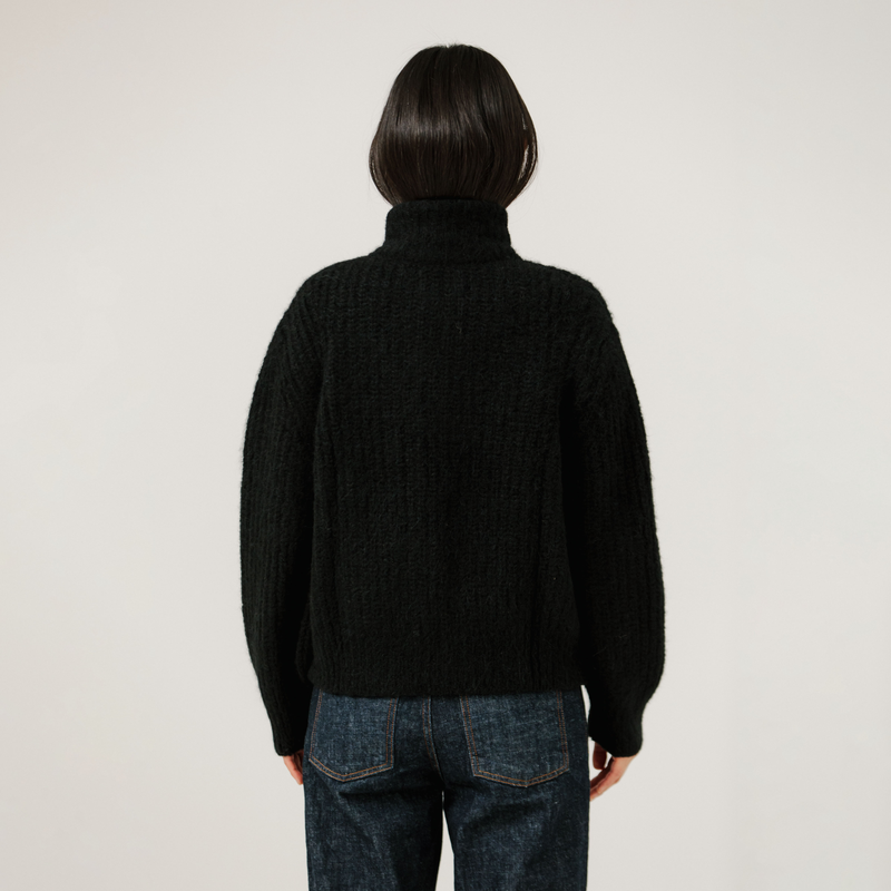 Person wearing a black turtleneck rib knit sweater and blue jeans on a plain background