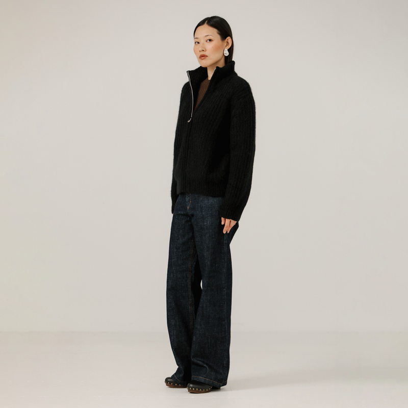 Person wearing a black zip up rib knit jacket and dark jeans on a plain background