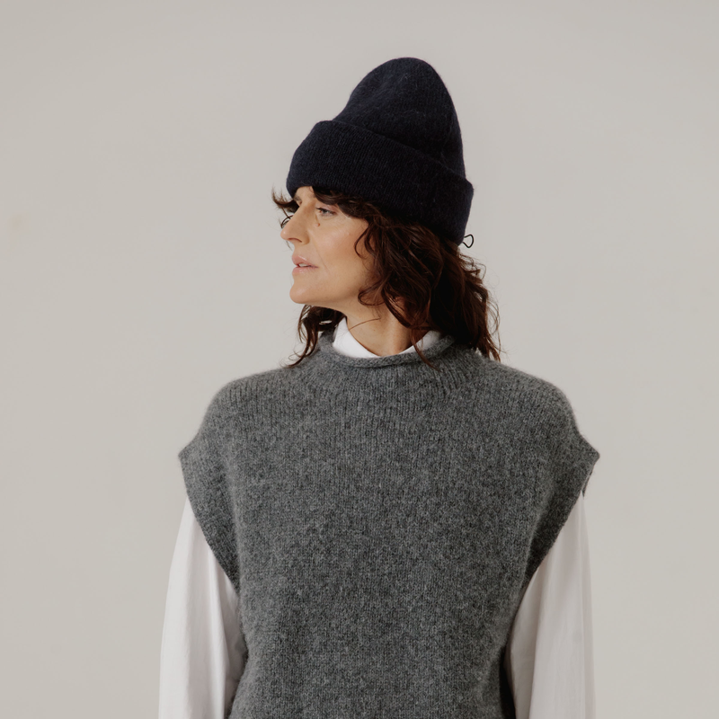 Woman wearing a dark blue beanie and gray sleeveless sweater against a plain background