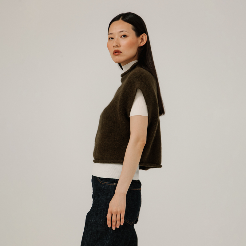 Woman wearing a brown vest over a white shirt with dark jeans on a plain background