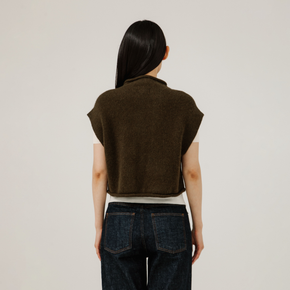 Person wearing a brown sleeveless sweater and dark jeans on a plain background
