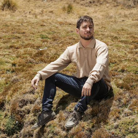Man sitting on a grassy hill wearing a beige shirt and dark pants.