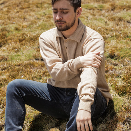 Man sitting on a grassy field wearing a beige sweater and dark pants.