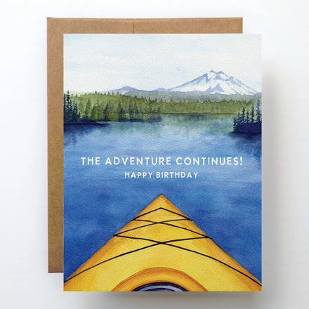 Greeting card with a kayak on water and mountain view, featuring 'The Adventure Continues! Happy Birthday' text.