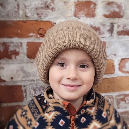 Child wearing a beige knit hat and patterned jacket against a brick wall.